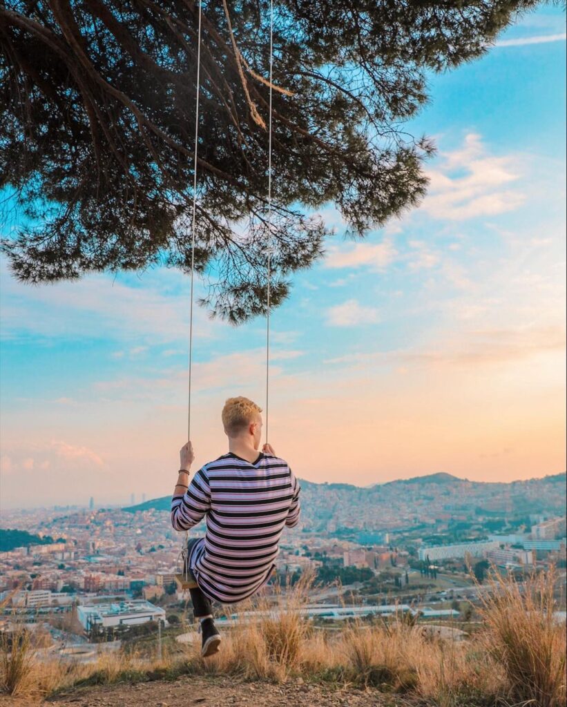 Young man on a swing with a black and white stripe shirt, swinging from a tree swing. In the distance we see the Barcelona cityscape.