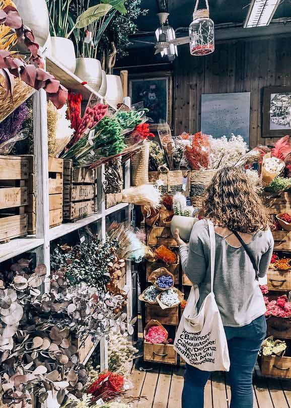 Young woman stands with her back against the camera looking at flowers. She is in a flower market, there are mix of flower bouquets in brown wooden boxes.